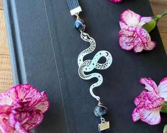 May include: A silver bookmark featuring a snake design with stars. The bookmark has two black beads with iridescent glitter. The bookmark is on a black book with pink flowers.
