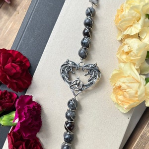 May include: A silver bookmark with two wolf heads forming a heart shape with a Celtic knot in the center. The bookmark is strung with gray and black beads and a black tassel.