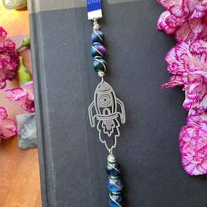 May include: A silver rocket ship bookmark with a blue tassel and iridescent blue and black beads. The bookmark is on a black book.