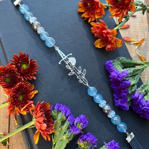 May include: A blue glass beaded bookmark with a silver sword charm and a black tassel. The bookmark is laying on a black book with orange and purple flowers.