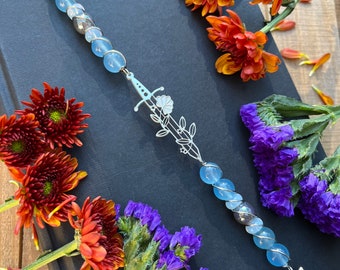 Peut inclure: Un marque-page en perles de verre bleu avec un charme d'épée argenté et un pompon noir. Le marque-page est posé sur un livre noir avec des fleurs orange et violettes.
