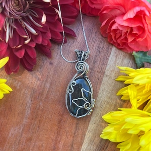 May include: A handcrafted silver pendant necklace featuring a teardrop-shaped, dark blue gemstone. The stone is encased in a wire design with floral accents and small gold-colored beads. The necklace is displayed with red and yellow flowers.