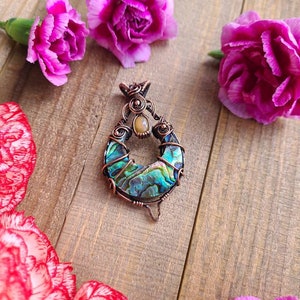 May include: A handcrafted pendant with a copper wire frame, featuring a vibrant abalone shell inlay. The pendant is surrounded by pink carnation flowers on a wooden surface. The abalone shell displays iridescent blues and greens.