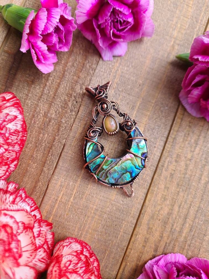 May include: A handcrafted copper pendant with an abalone shell inlay, featuring iridescent blues, greens, and purples. The pendant is accented with a small, round, yellow gemstone. Pink carnations and a wooden surface provide the background.