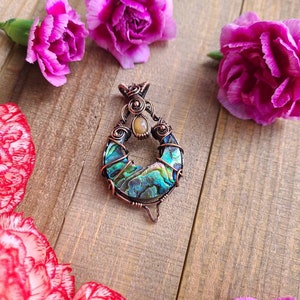 May include: A handcrafted pendant with a teardrop shape, featuring iridescent abalone shell and copper wire detailing. The pendant is adorned with a small, oval-shaped stone. The background includes pink carnation flowers on a wooden surface.