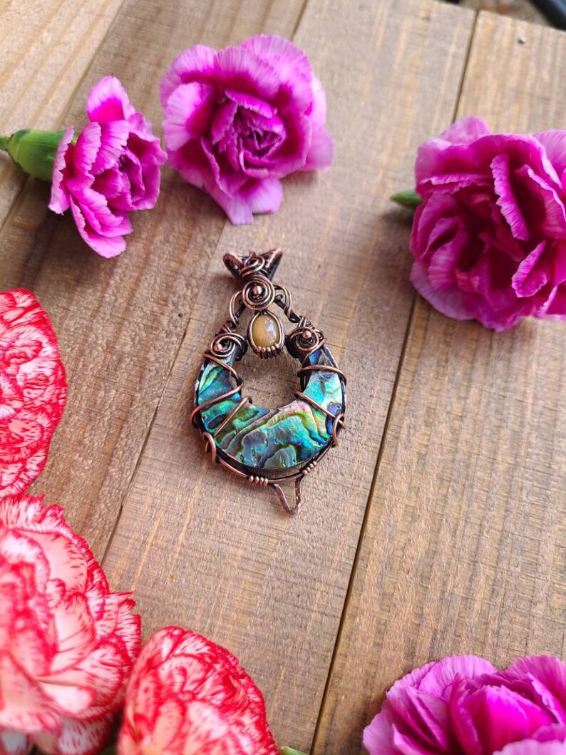 May include: A handcrafted pendant with a teardrop shape, featuring iridescent abalone shell and copper wire detailing. The pendant is adorned with a small, oval-shaped stone. The background includes pink carnation flowers on a wooden surface.