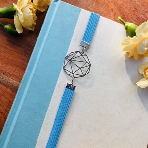 Könnte beinhalten: Ein hellblaues Lesezeichen mit einem geometrischen silbernen Anhänger. Das Lesezeichen befindet sich auf einem blauen und weißen Buch.