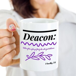 May include: White ceramic mug with black text that reads "Deacon: holding the mystery of the faith with great boldness 1 Timothy 3:5". The text is in a purple wavy line with a purple leaf design.