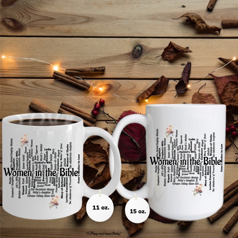 May include: Two white ceramic mugs with black handles. The mugs feature a black and white word cloud design with the text "Women in the Bible". The mugs are on a wooden surface with a string of lights and fall leaves. The mugs are available in 11 oz and 15 oz sizes.