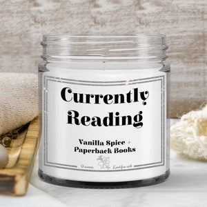 May include: A white candle jar with a silver glitter label that reads "Currently Reading" and "Vanilla Spice + Paperback Books". The label also includes the text "9 ounces" and "Lead-free wick".