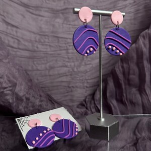 Pode incluir: Um par de brincos de declaração roxo e rosa com um design ondulado. Os brincos são exibidos em um suporte prateado.