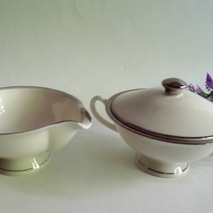 May include: Cream-colored serving set with silver trim. The set includes a gravy boat with a handle and a covered sugar bowl. Both pieces have a pedestal base and a silver accent around the rim.
