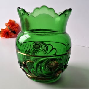 May include: A vibrant green glass vase with a scalloped rim and ornate gold accents. The vase features detailed embossed floral designs and a gold band around the middle. Decorative orange flowers are in the background.