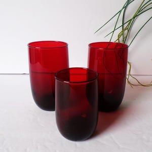 May include: Three red-tinted drinking glasses. The glasses have a rounded, cylindrical shape with a darker base. The glasses are made of a transparent material, allowing light to pass through. The background is white.