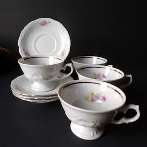 May include: A set of white porcelain teacups and saucers. The cups have a pedestal base and ornate handles. Each piece is decorated with a gold rim and a floral design in shades of purple, yellow, and pink. The saucers have a scalloped edge.