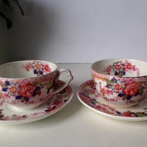 May include: Two matching teacups and saucers with floral patterns. The cups and saucers are white with a pink border and feature a design of red, pink, blue, and green flowers. The cups have a curved handle.