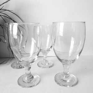 May include: A set of four clear, stemmed wine glasses. The glasses have a rounded bowl shape and sit on a short stem with a circular base. The glasses are arranged on a white surface, with a plant in the background.