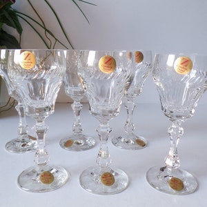 May include: Set of six clear crystal wine glasses. Each glass has a round base, a faceted stem, and a bowl with a textured, swirling design. Gold foil stickers with text are affixed to each glass. The glasses are arranged on a white surface.