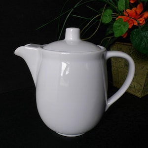 May include: A white ceramic teapot with a lid and a curved handle. The teapot has a spout for pouring and a small knob on top of the lid. The teapot is sitting on a black surface.