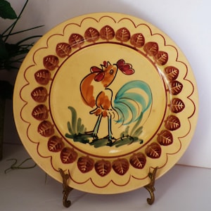 May include: A vintage ceramic plate with a rooster illustration. The plate features a yellow center with a brown and orange rooster, blue tail feathers, and green grass. The rim has a scalloped edge with brown leaf designs. The plate is displayed on a gold-colored stand.