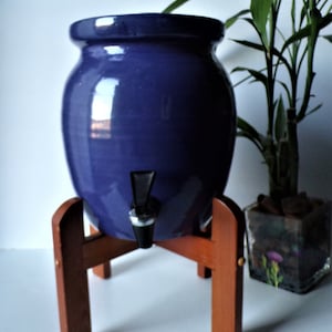May include: A deep blue ceramic beverage dispenser with a spigot, set on a wooden stand. The dispenser has a rounded shape and a glossy finish. A bamboo plant in a glass cube is in the background.