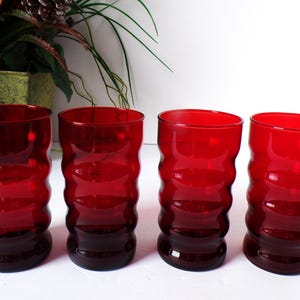 May include: Four red glass tumblers with a wavy, textured design. The glasses have a deep red color that fades to a darker shade at the base. The glasses are cylindrical with a wide opening.