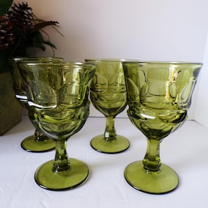 May include: Four olive green vintage goblets with a textured pattern. The goblets have a rounded bowl shape and a stem with a circular base. The goblets are arranged on a white surface, with a decorative plant in the background.