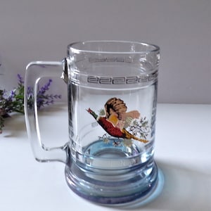 May include: A clear glass beer mug featuring a colorful illustration of a pheasant in flight. The mug has a sturdy handle and a decorative Greek key pattern etched around the upper rim. The base is thick and slightly tinted with a blue hue.
