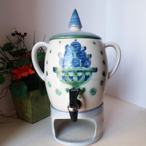 May include: A ceramic beverage dispenser with a white base and a blue and green floral design. The dispenser has a spigot, two handles, and a lid with a pointed top. The design includes blue berries in a green bowl.