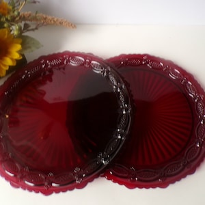 May include: Two ruby red glass plates with a scalloped edge and a detailed border. The plates have a radial design and are likely vintage or antique. The plates are round and are a deep, rich red color.