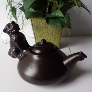 May include: A dark brown ceramic teapot with a lid and a sculpted animal figure on the side. The teapot has a rounded body and a spout. The background includes a decorative green square vase with a floral arrangement.