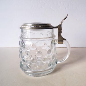 May include: Clear glass beer stein with a silver-colored metal lid and handle. The body of the stein has a textured, bubble-like pattern. The lid is hinged and has a decorative edge.