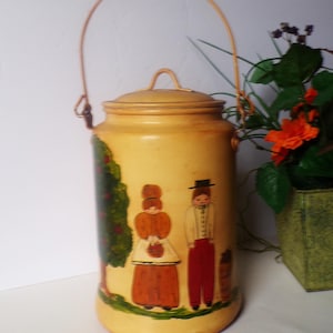 May include: A cylindrical yellow metal canister with a matching lid and a wire handle. The exterior features a hand-painted scene of a green apple tree and two figures standing on a patch of grass, wearing orange and red clothing.