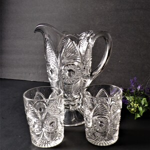 May include: A clear glass pitcher and two matching glasses. The pitcher has a handle and a spout. The glasses and pitcher have a detailed, embossed pattern. The set is likely for serving beverages.