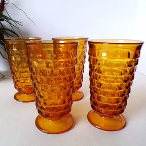 May include: Four amber-colored drinking glasses. Each glass has a textured, stacked brick pattern and a rounded base. The glasses are made of glass and are suitable for serving beverages. The glasses are arranged on a white surface.