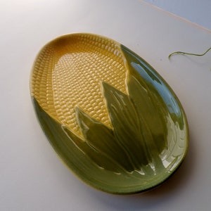 May include: Oval-shaped ceramic serving dish with a corn-on-the-cob design. The dish features a textured yellow center, mimicking corn kernels, and is framed by green leaves. The outer rim is also green.