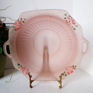 May include: A vintage pink glass serving tray with a scalloped edge and two handles. The tray features a central raised section and is decorated with small pink floral accents. It is displayed on a decorative gold-colored stand.