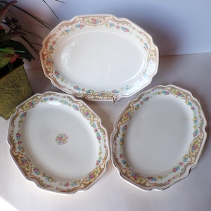 May include: Three oval-shaped ceramic platters with scalloped edges. Each piece features a cream-colored center and a decorative border with a repeating pattern of small pink, blue, and yellow flowers on a tan background with gold trim.