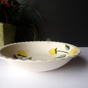 May include: Off-white ceramic serving bowl with a scalloped edge. The bowl is decorated with yellow flower and green leaf designs. The bowl is likely vintage and is suitable for serving food.