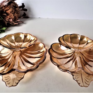 May include: Two iridescent, amber-colored glass serving dishes. Each dish has a three-sectioned design with a scalloped edge and a leaf-shaped base. The dishes are displayed on a white surface, with dried flowers and greenery in the background.