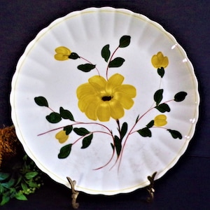 May include: A round white plate with a scalloped edge featuring a hand-painted design of a large central yellow flower with dark centers, surrounded by smaller yellow buds and dark green leaves on thin reddish-brown stems against a white background.