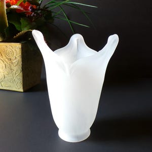 May include: A white, frosted glass vase with a tulip-like shape. The vase has a wide, open top with curved edges and a rounded base. The smooth, opaque surface reflects light, creating a soft, elegant appearance. This decorative vase is suitable for floral arrangements.