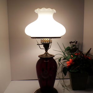 May include: A table lamp featuring a deep red glass base with a floral pattern and a brass-colored metal neck. The lamp is topped with a white, ruffled-edge glass shade that emits a soft glow, supported by a decorative brass-toned metal gallery.