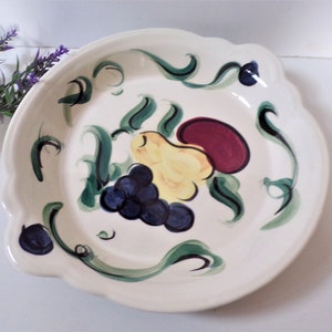 May include: Off-white ceramic serving dish with a scalloped edge. The dish features a hand-painted design of a bunch of dark blue grapes, a yellow pear, and a maroon bean-shaped fruit. Green vines and leaves decorate the rim.