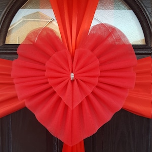 May include: A red tulle bow in the shape of a heart is tied to a red satin ribbon draped over a black door. The bow has a small, sparkly rhinestone in the centre.