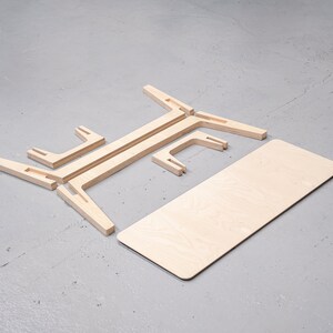Puede incluir: Una mesa de madera de paquete plano con un acabado de madera clara. La mesa est&aacute; desmontada y colocada sobre una superficie gris. La superficie de la mesa es rectangular y las patas est&aacute;n hechas de cuatro piezas de madera que encajan entre s&iacute;.