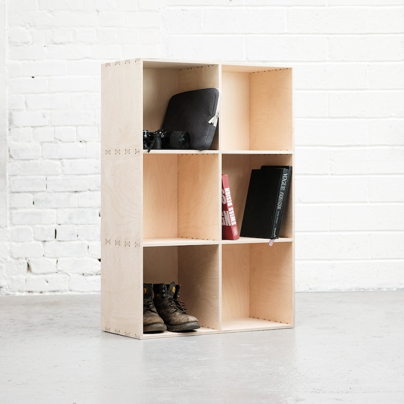 May include: A light wood cube shelf unit with six compartments. The top shelf has a black laptop case, the middle shelf has a red book titled "Swiss Design" and a black book titled "Vogue de Maison", and the bottom shelf has two brown work boots.