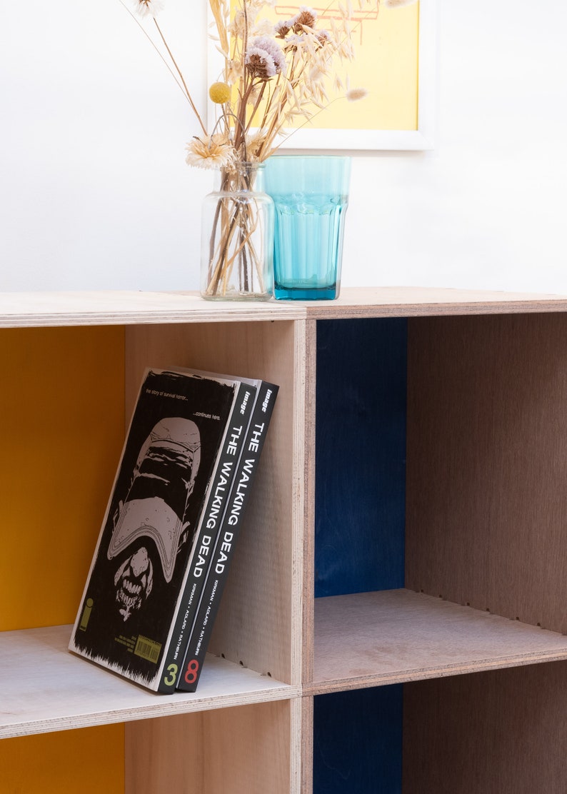May include: A wooden shelf unit with three shelves, painted in yellow, blue, and natural wood. Two graphic novels titled "The Walking Dead" are on the shelf.