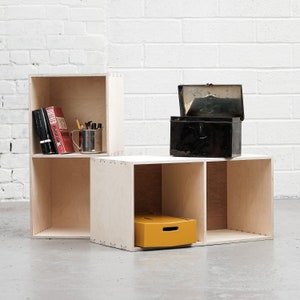 May include: A set of three wooden cube shelves with open compartments. The shelves are made of light wood and have a natural finish. The top shelf has a red book, a silver cup, and pencils. The middle shelf is empty. The bottom shelf has a yellow box. A black metal box with a lid is on top of the shelves.