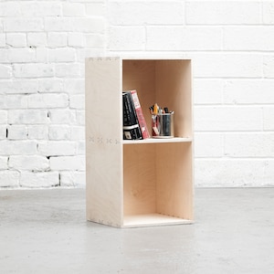 May include: A two-shelf wooden bookcase with a light wood finish. The top shelf has a few books and a metal cup with pencils. The bottom shelf is empty.
